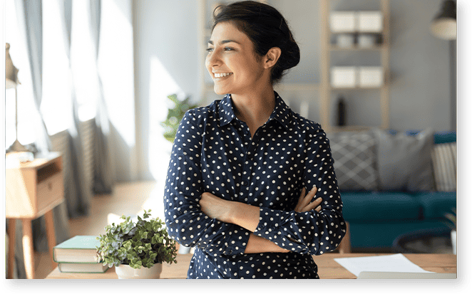 A woman in a navy blue polka dot shirt stands with folded arms, smiling and looking to the side in a well-lit room, reflecting the expertise of lighting analysts support, with shelves and a couch in the background.