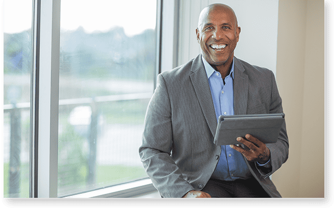 Smiling man in a suit sits by a window holding a tablet, ready to offer lighting analysts support.